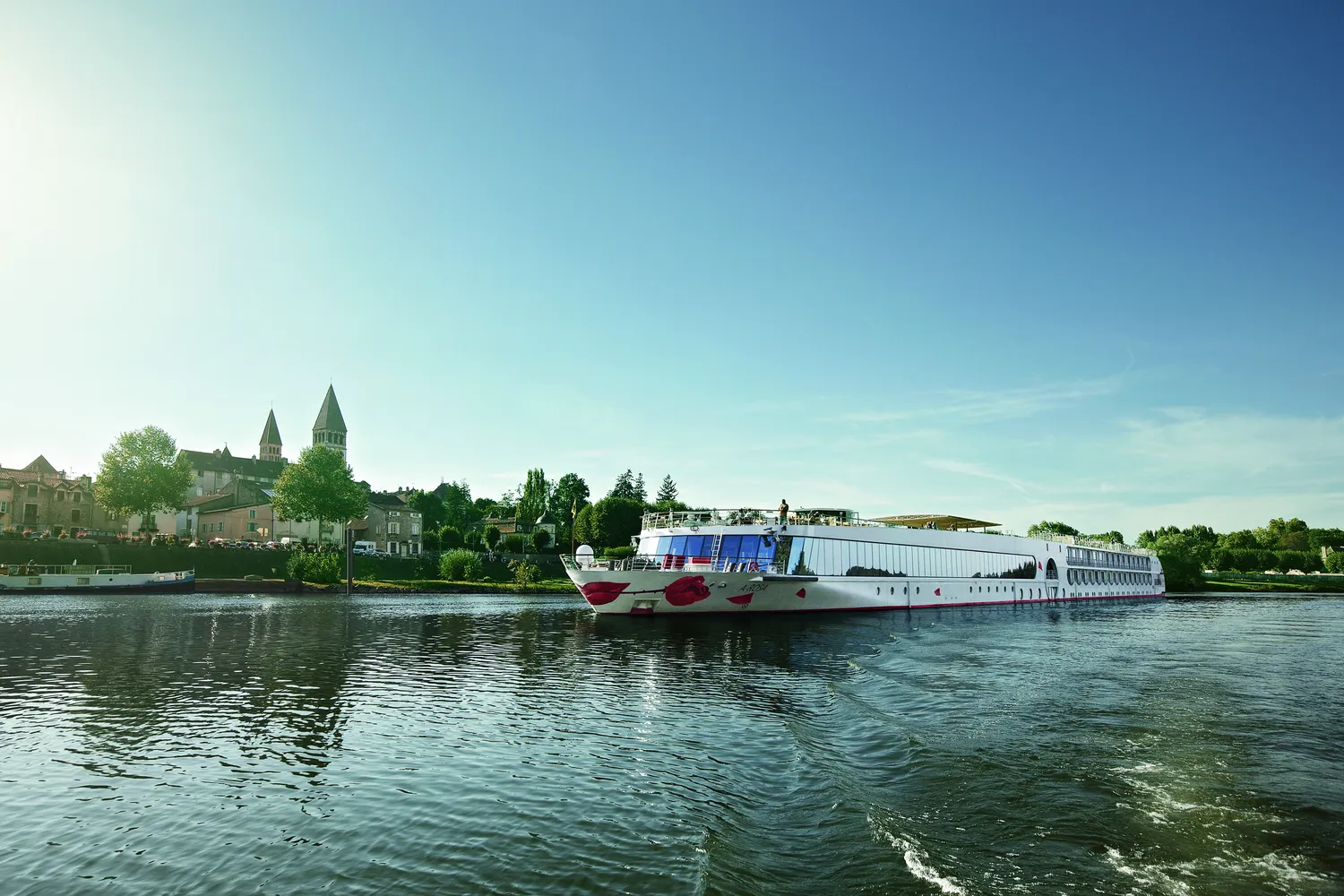 GTS Reiseservice – Ein elegantes weißes Flusskreuzfahrtschiff, verziert mit einer roten Lippe und einer Rose, gleitet ruhig auf einem Fluss vorbei an einer Küstenstadt mit markanten Kirchtürmen.