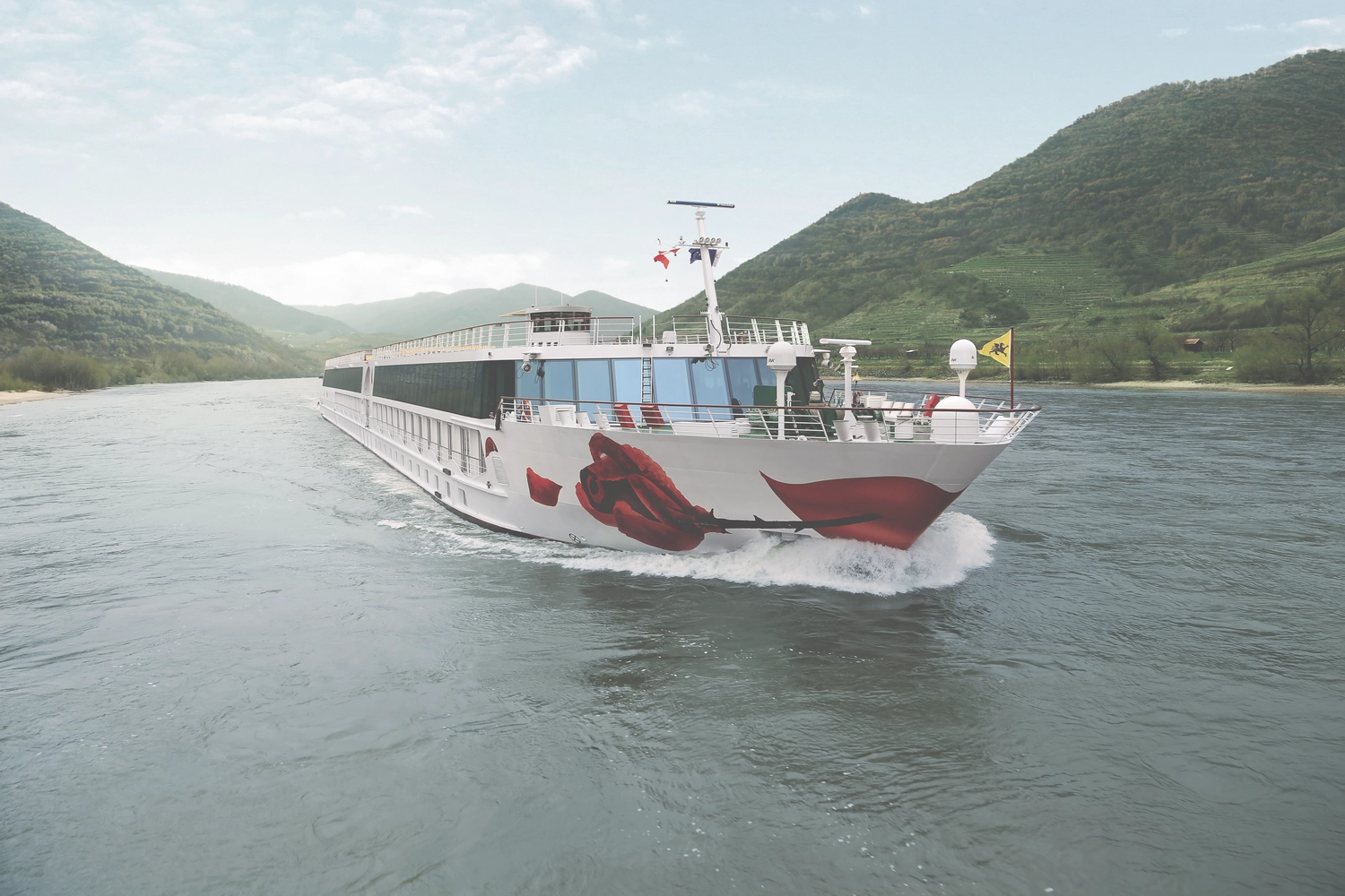GTS – Reiseservice – Ein weißes Flusskreuzfahrtschiff mit einer großen roten Rose fährt auf einem Fluss, der von grünen Hügeln gesäumt ist.