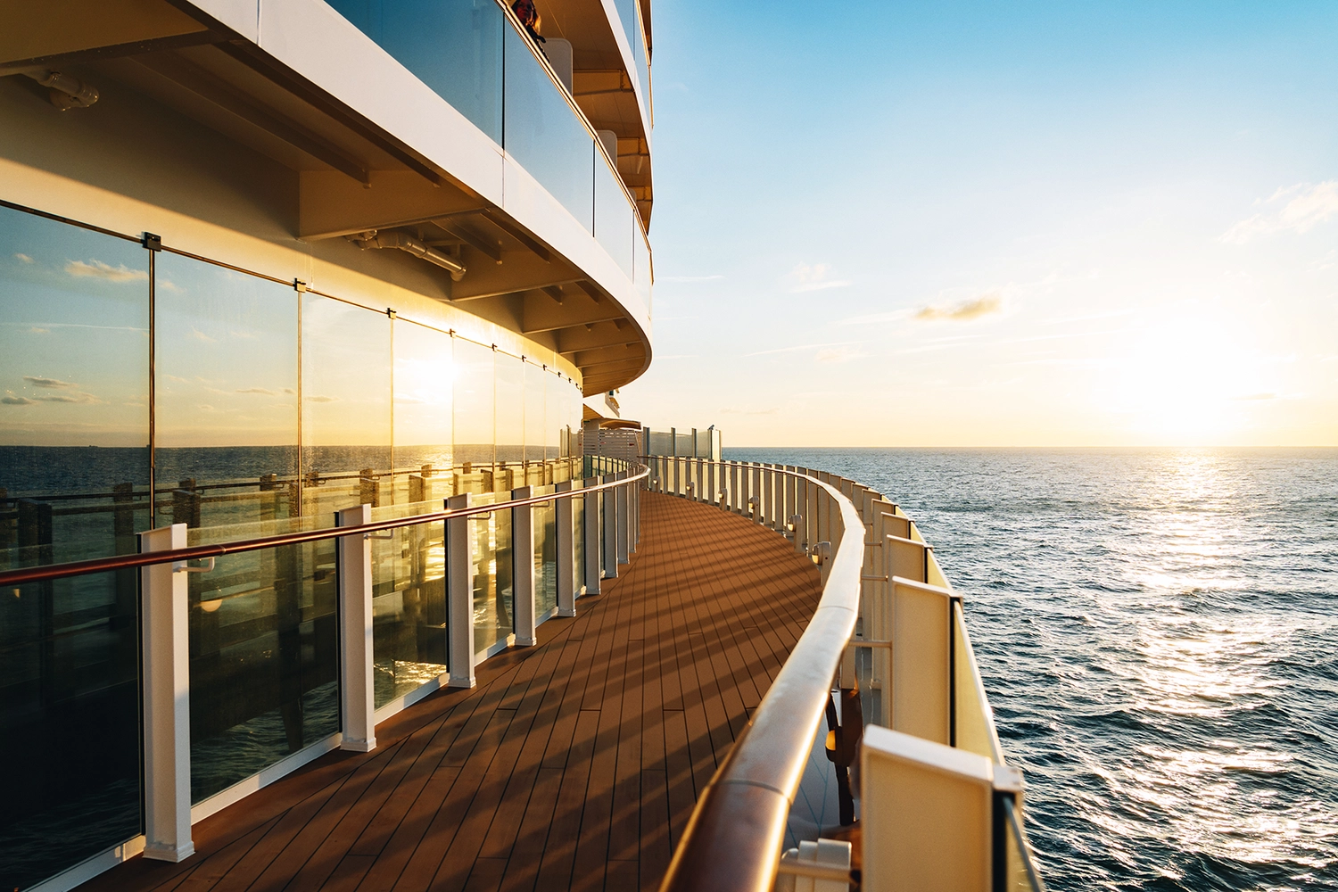 GTS – Reiseservice – Das geschwungene Holzdeck eines Kreuzfahrtschiffs im Sonnenuntergang, dessen Glasgeländer das goldene Licht des Ozeans reflektieren.