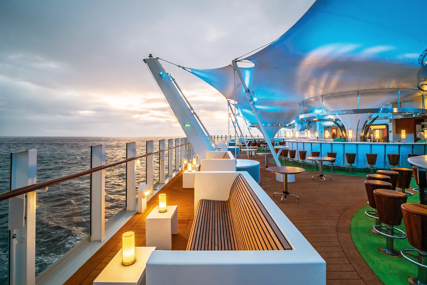 GTS - Reiseservice - Eine moderne Decklounge auf einem Kreuzfahrtschiff, die im Sonnenuntergang in leuchtendem Blau erstrahlt.