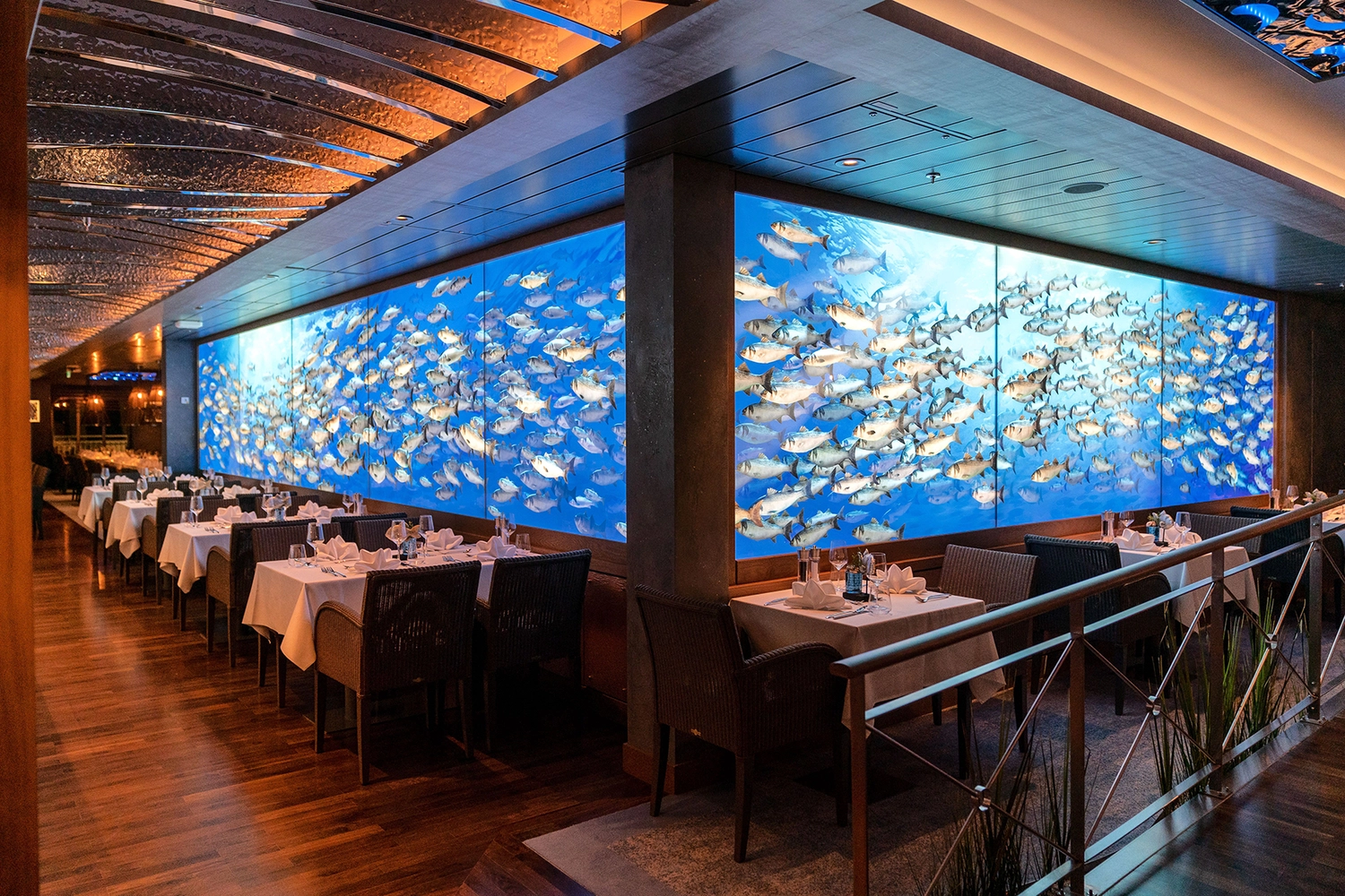 GTS - Reiseservice - Das Restaurant auf dem Kreuzfahrtschiff mit gedeckten Tischen, die eine große Aquariumwand mit vielen Fischen flankieren.