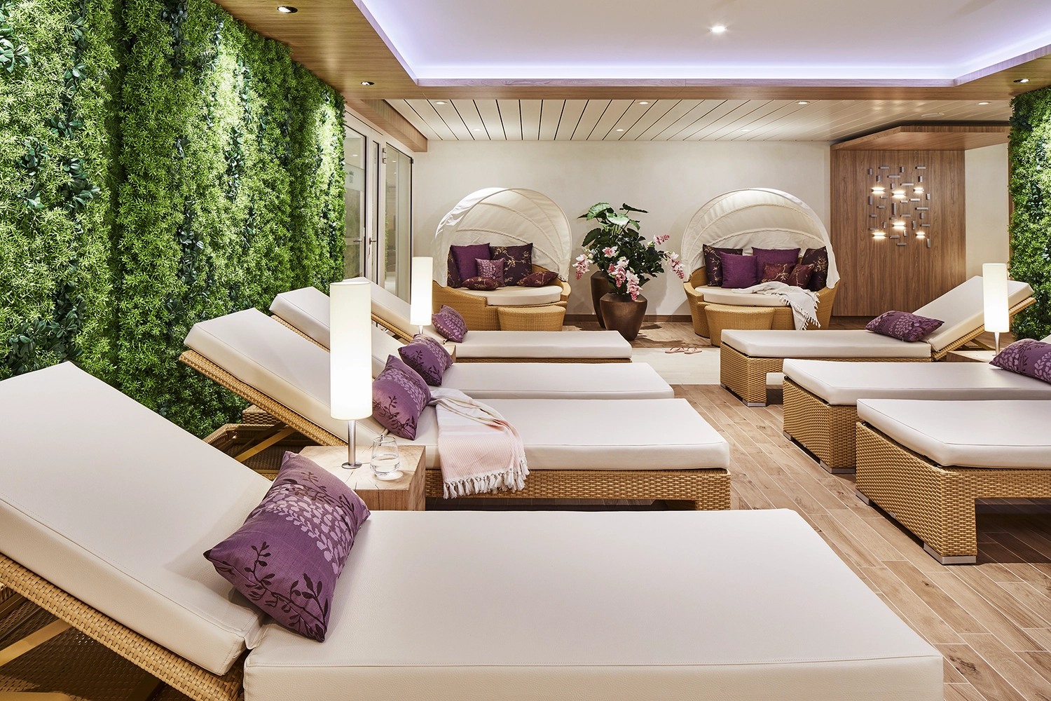 GTS - Reiseservice - Entspannungsraum im Spa eines Kreuzfahrtschiffs mit Korbsesseln, violetten Kissen und einer üppig begrünten Wand.