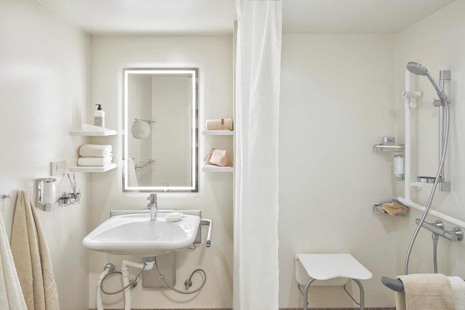 GTS - Reiseservice - Ein barrierefreies, modernes Badezimmer mit Waschbecken mit beleuchtetem Spiegel und ebenerdiger Dusche mit Sitzgelegenheit.