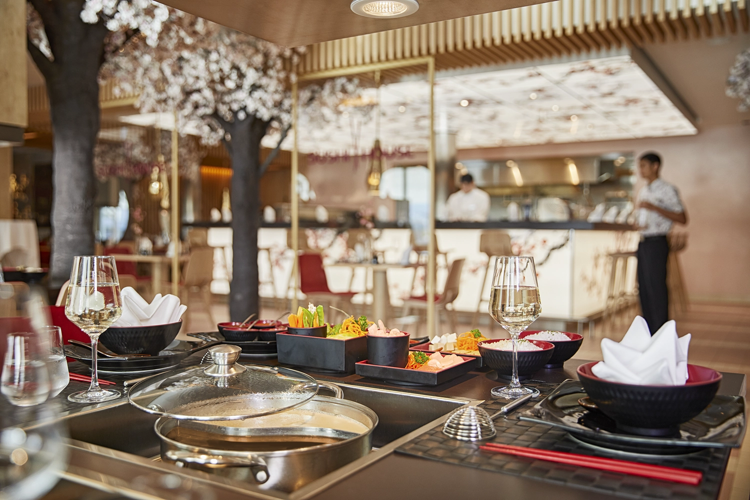 GTS - Reiseservice - Ein gedeckter Tisch mit Hot Pot, Beilagen und Weingläsern in einem modernen Restaurant, das mit Kirschblütenbäumen dekoriert ist.