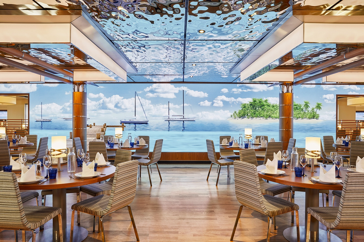 GTS – Reiseservice – Ein helles Kreuzfahrtschiffrestaurant mit runden Tischen und großen Fenstern, die den Blick auf Segelboote, einen Holzsteg und eine tropische Insel freigeben.