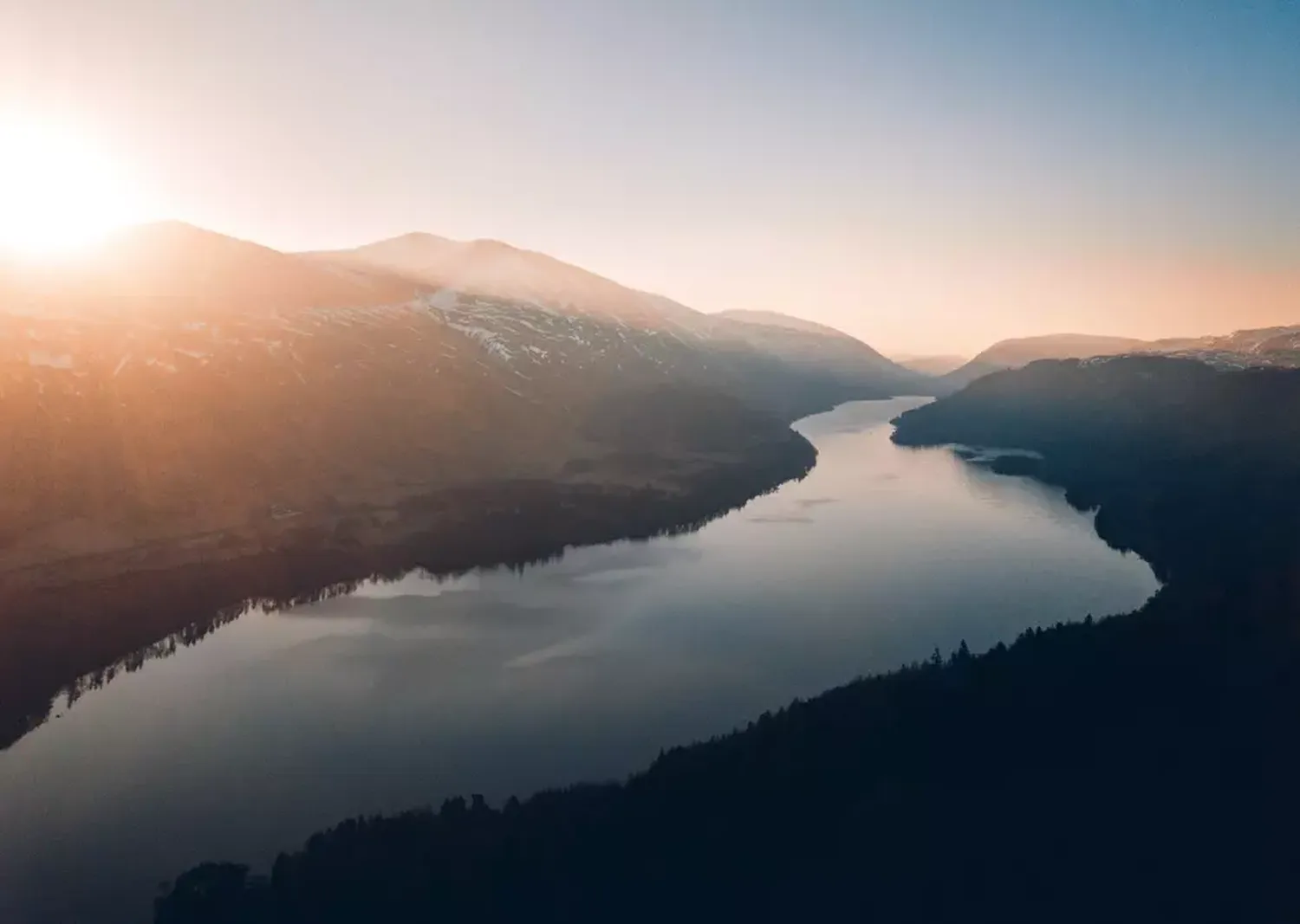 GTS - Reiseservice - Luftaufnahme eines breiten Flusses, der sich bei hellem, dunstigen Sonnenaufgang durch ein von schneebedeckten Bergen gesäumtes Tal schlängelt.