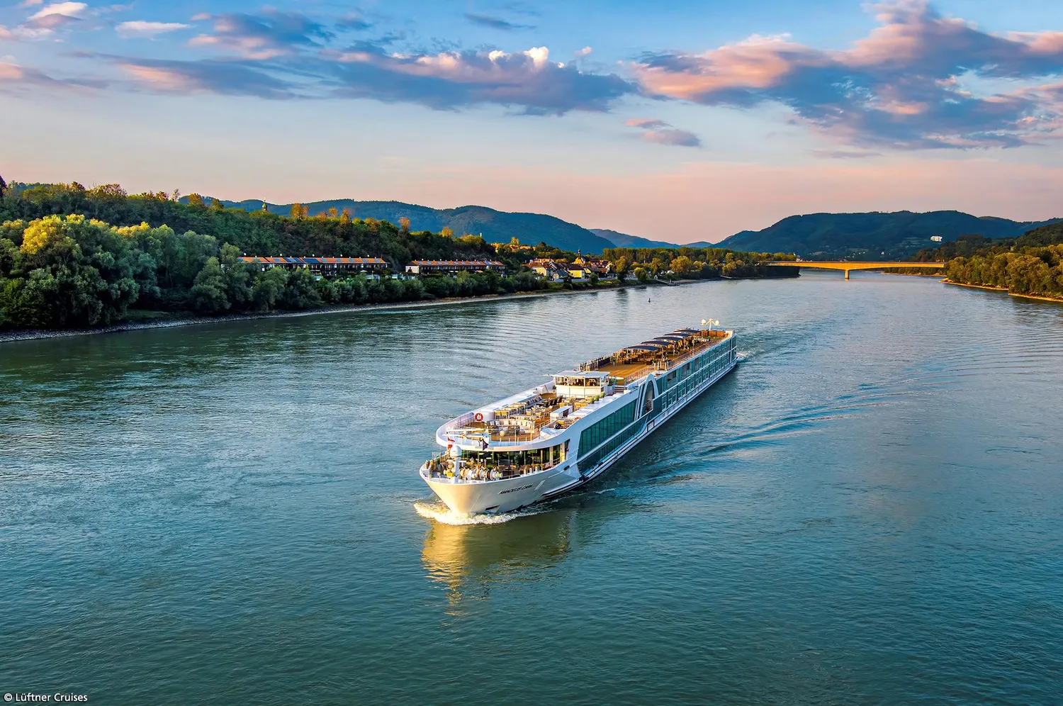 GTS - Reiseservice - Luftaufnahme eines langen Flusskreuzfahrtschiffs, das während eines warmen, goldenen Sonnenuntergangs einen breiten, von dichten Wäldern gesäumten Wasserweg befährt.