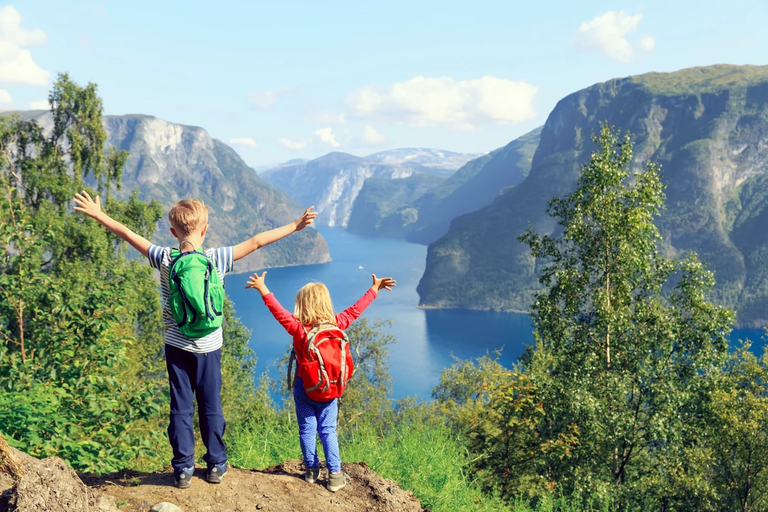 GTS - Reiseservice - Zwei kleine Kinder mit Rucksäcken stehen mit erhobenen Armen auf einem Aussichtspunkt und blicken auf einen weiten, malerischen Gebirgsfjord.