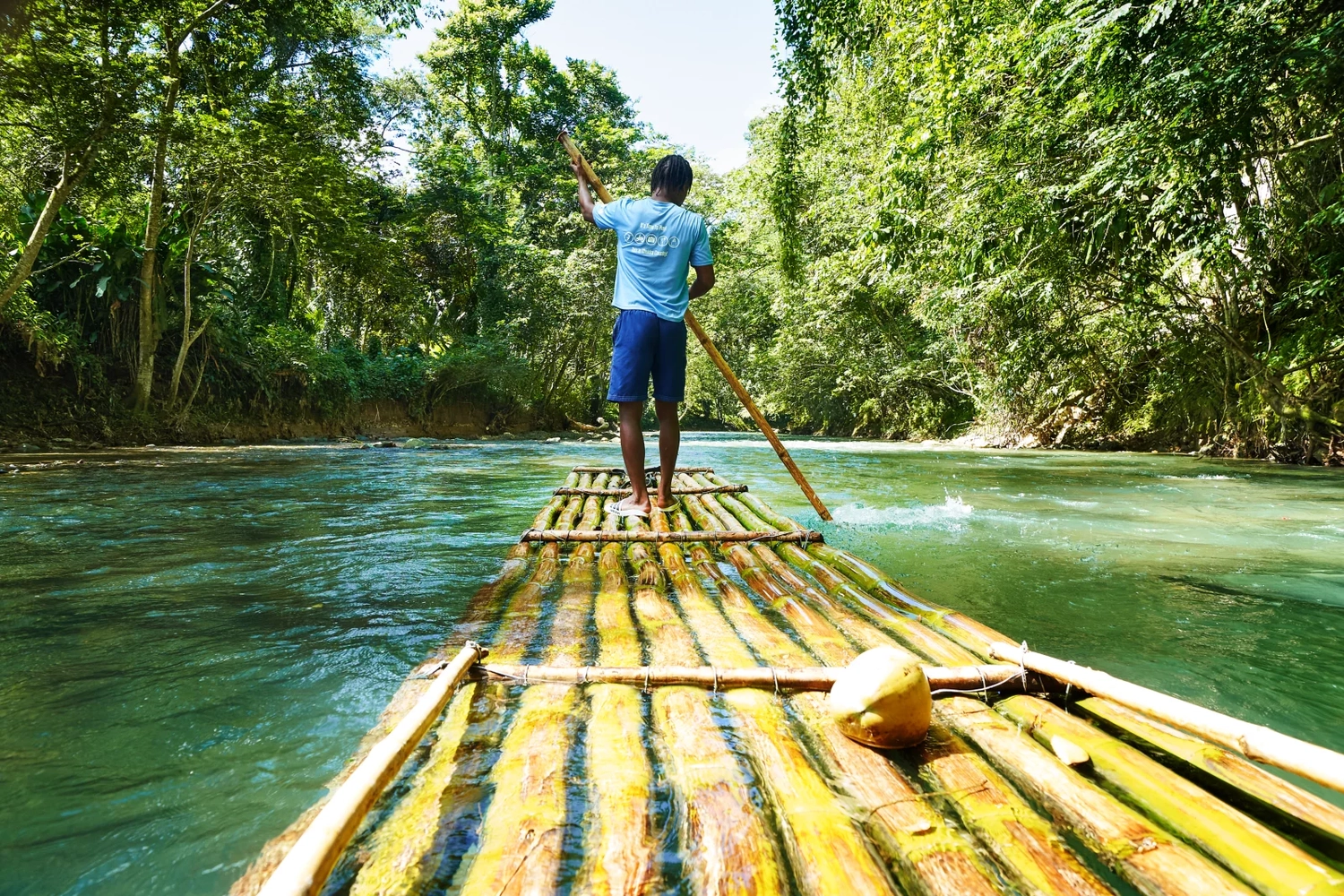 GTS - Reiseservice - Ein Mann steuert ein Bambusfloß auf einem tropischen Fluss, umgeben von üppigem, grünem Wald.