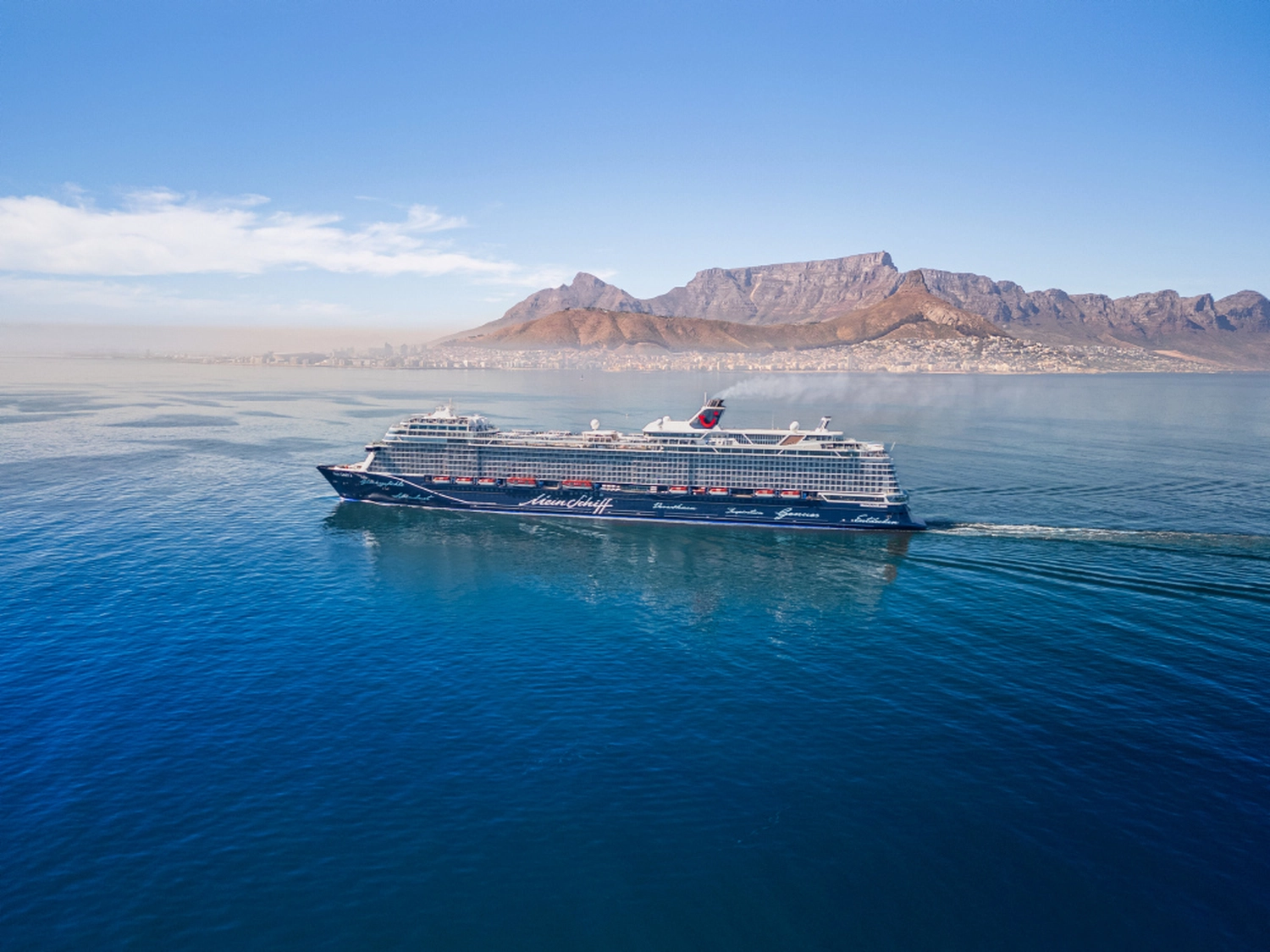 GTS - Reiseservice - Ein dunkelblaues Kreuzfahrtschiff, das über den Ozean navigiert, im Hintergrund eine markante, flache Bergkette.