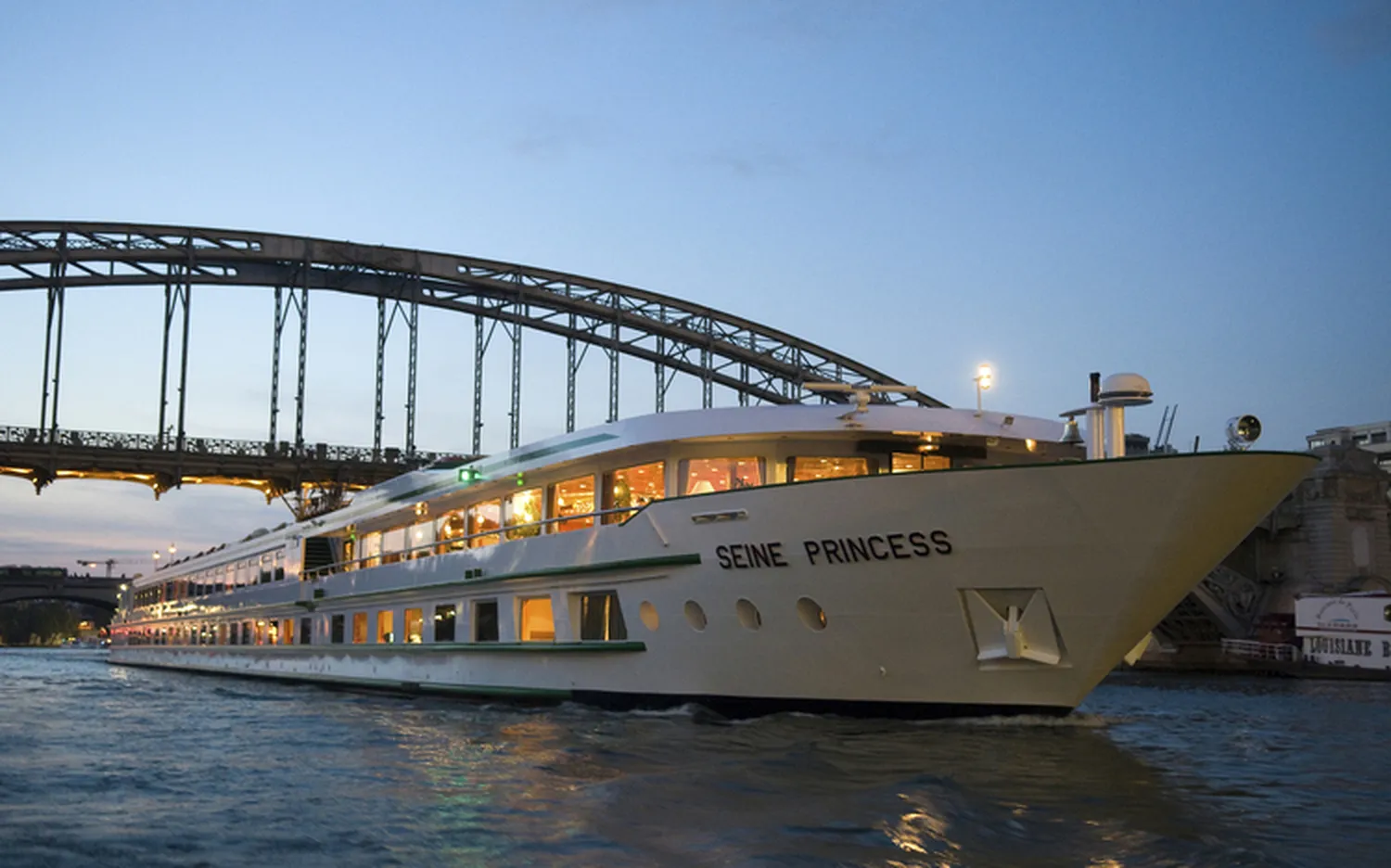 GTS – Reiseservice – Ein großes weißes Flusskreuzfahrtschiff namens „SEINE PRINCESS“ mit beleuchteten Fenstern fährt in der Abenddämmerung unter einer großen Stahlbogenbrücke hindurch.