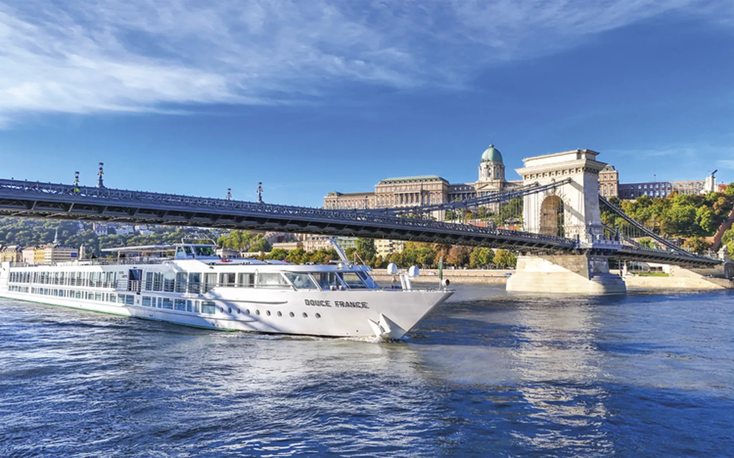 GTS – Reiseservice – Ein großes weißes Flusskreuzfahrtschiff namens „DOUCE FRANCE“ fährt unter einer historischen Hängebrücke hindurch. Im Hintergrund erhebt sich ein prächtiger Palast mit Kuppel am Hang.