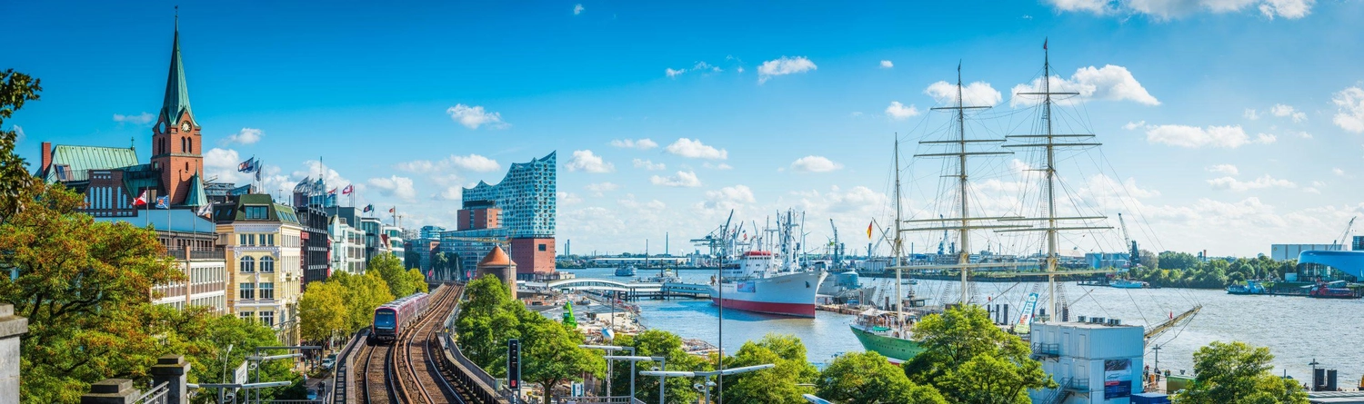 GTS - Reiseservice - Hafenstadtpanorama mit Schiffen, Bahngleisen, einem historischen Kirchturm und moderner Architektur unter strahlend blauem Himmel.