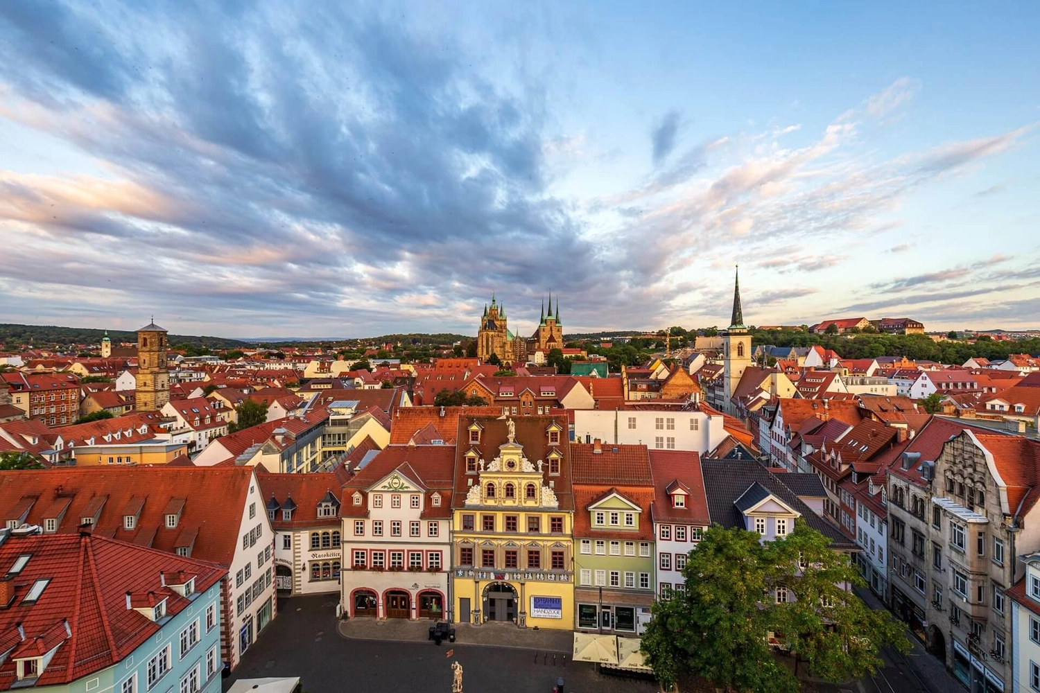 GTS – Reiseservice – Blick aus der Vogelperspektive auf einen historischen Stadtplatz, umgeben von Gebäuden mit roten Dächern und Kirchtürmen in der Ferne, unter einem bewölkten Abendhimmel.