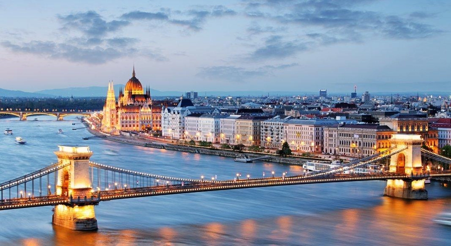 GTS - Reiseservice - Beleuchtete Hängebrücke und das imposante Parlamentsgebäude an einer Flussstadt in der Abenddämmerung.