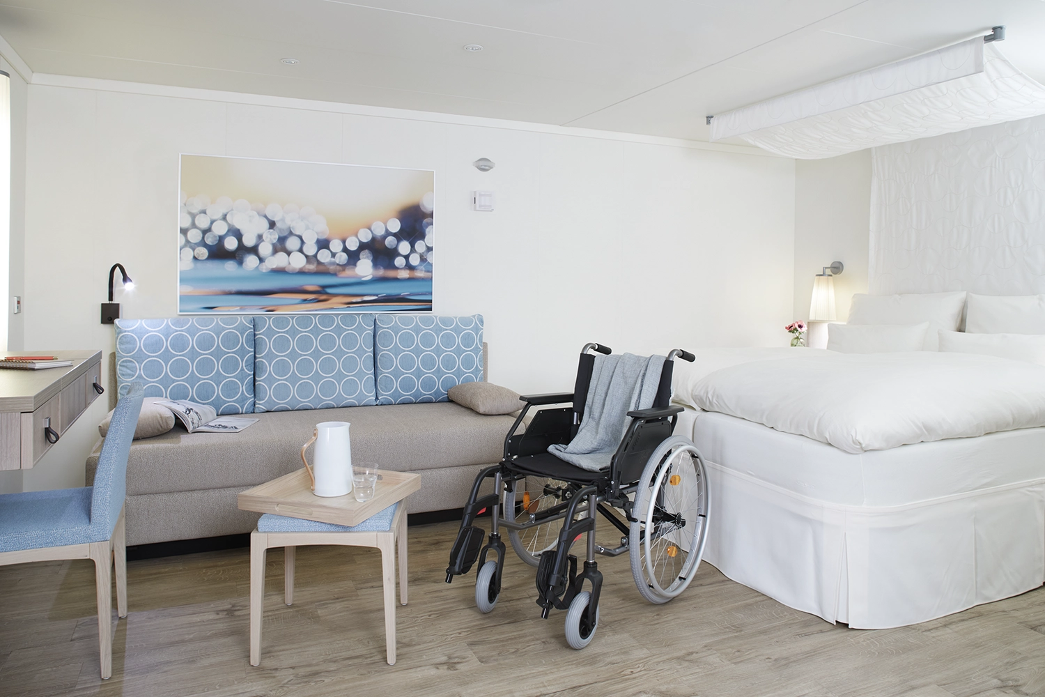GTS - Reiseservice - Ein barrierefreies Hotelzimmer mit einem großen weißen Bett, einem Sofa und einem leeren Rollstuhl in der Mitte.