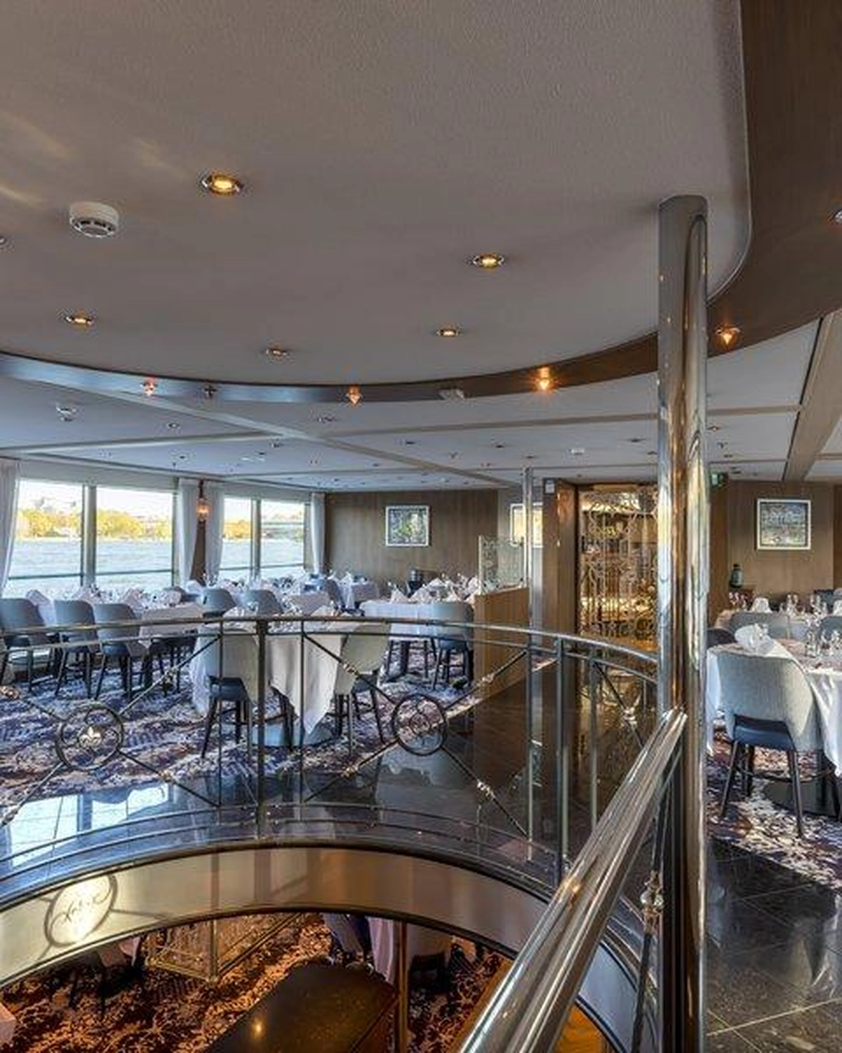 GTS - Reiseservice - Mehrstöckiger Speisesaal eines Kreuzfahrtschiffs mit weiß gedeckten Tischen und großen Fenstern mit Flussblick.