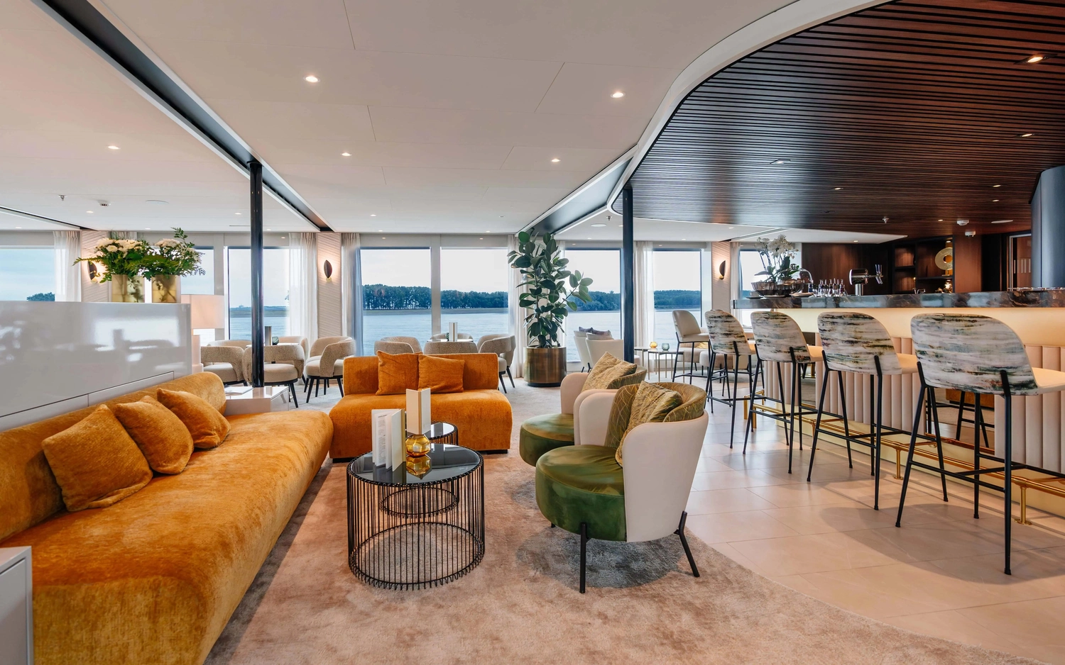 GTS – Reiseservice – Ein moderner Loungebereich auf einem Kreuzfahrtschiff mit bequemen orangefarbenen Sofas, grünen Samtsesseln und einer eleganten Bar mit Barhockern und Blick auf den Fluss.