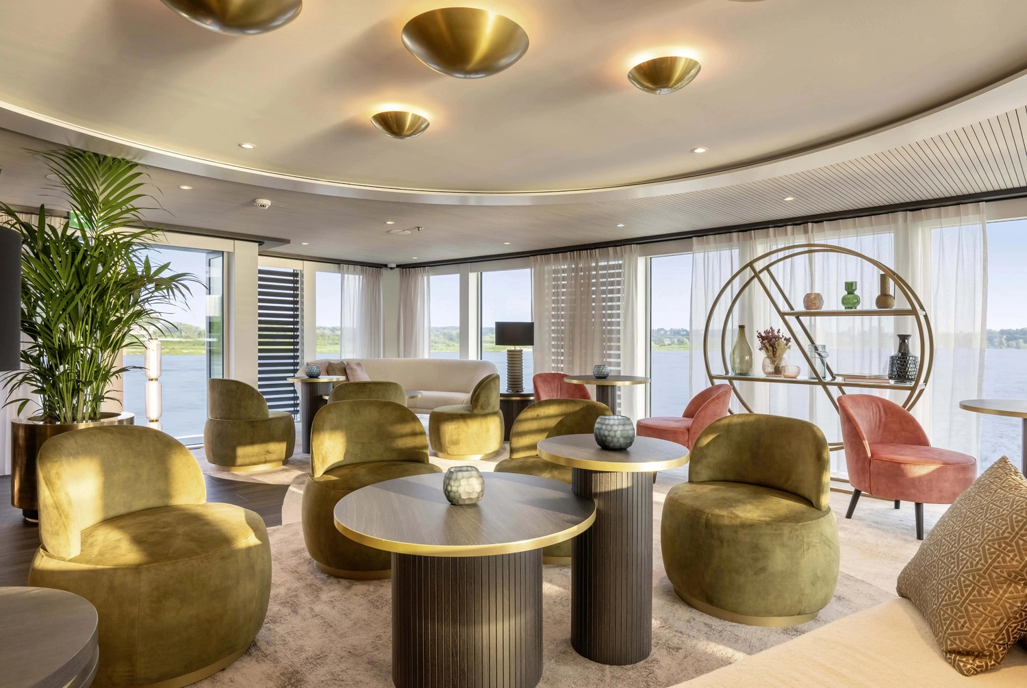 GTS - Reiseservice - Moderne Kreuzfahrtlounge mit olivgrünen und rosafarbenen Sesseln, runden Tischen und großen Fenstern mit Flussblick.