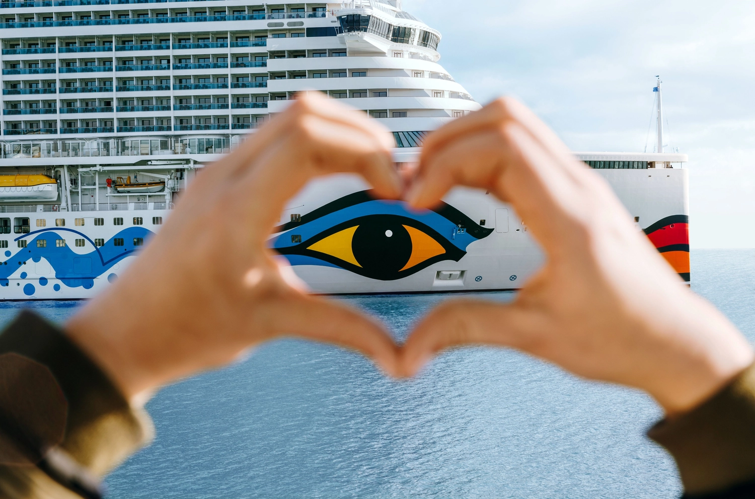 GTS - Reiseservice - Hände formen ein Herz um ein großes Kreuzfahrtschiff, das mit einem auffälligen Augenmotiv verziert ist.