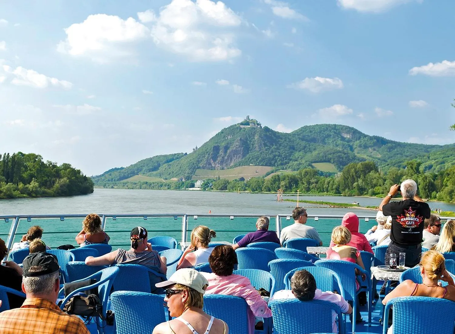 GTS - Reiseservice - Passagiere entspannen sich auf blauen Stühlen an Deck eines Flussschiffs und blicken auf einen markanten grünen Hügel mit einem Gebäude auf der Spitze.